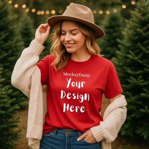 Puede incluir: Una camiseta roja con el texto "Your Design Here" en blanco, usada con vaqueros azules y un sombrero marrón. La camiseta también tiene el texto "MocksyDaisy" encima del texto principal. Un cárdigan beige está drapeado sobre los hombros.
