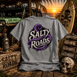 May include: A heather gray short-sleeve t-shirt featuring a circular graphic on the back. The design includes a purple octopus tentacle wrapped around the white text SALTY ROADS. The shirt is displayed against a backdrop of a vehicle and a surfboard.