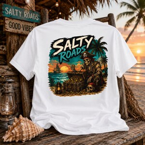 Puede incluir: Una camiseta blanca con un estampado gr&aacute;fico de un esqueleto pirata sentado en una playa junto a un cofre del tesoro, un farol y una palmera al atardecer. El texto "SALTY ROADS" est&aacute; impreso en una fuente estilizada sobre la ilustraci&oacute;n.