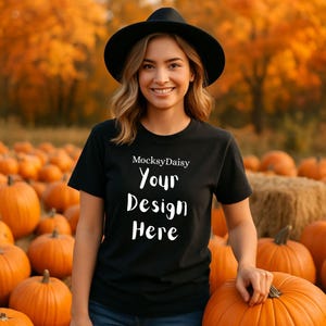 May include: A black t-shirt with the text "MocksyDaisy Your Design Here" in white. The person is wearing a black hat and is standing in a pumpkin patch. The background features orange pumpkins and autumn foliage.