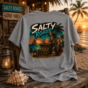 Puede incluir: Una camiseta gris jaspeado con un estampado gr&aacute;fico de un esqueleto con sombrero de pirata, sentado junto a un cofre del tesoro en una playa al atardecer. El texto SALTY ROADS aparece en letras grandes sobre el esqueleto, con palmeras.