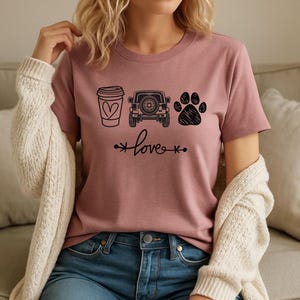 May include: Mauve-colored t-shirt featuring black graphic icons of a coffee cup with a heart, a jeep, and a paw print. The word "love" is written in cursive below the icons, with arrows on each side.