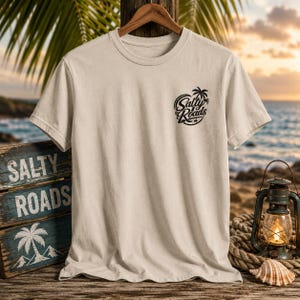 May include: A beige short-sleeve t-shirt featuring a black graphic on the left chest that displays the text Salty Roads with a palm tree icon. The shirt is displayed on a wooden hanger against a backdrop of a beach scene with a lantern and sign.
