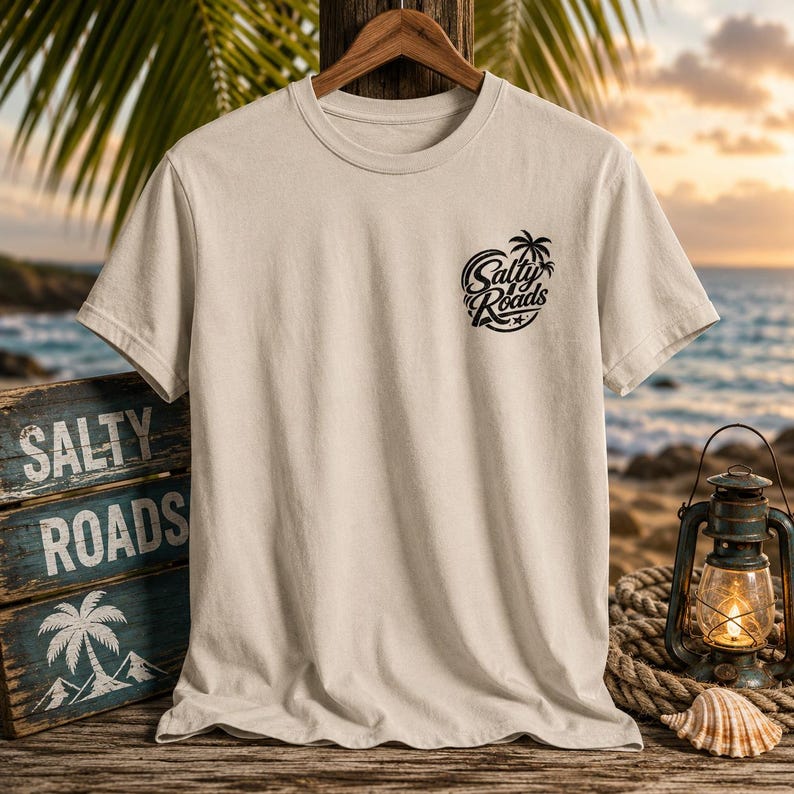 May include: A beige short-sleeve t-shirt featuring a black graphic on the left chest that displays the text Salty Roads with a palm tree icon. The shirt is displayed on a wooden hanger against a backdrop of a beach scene with a lantern and sign.