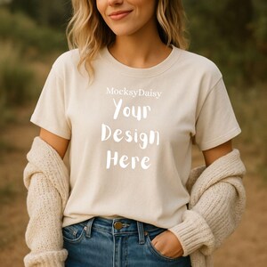 Könnte beinhalten: Ein cremefarbenes T-Shirt mit dem weißen Text "Your Design Here" und dem Markennamen "MocksyDaisy" darüber. Das Model trägt eine blaue Jeans und einen beigen Cardigan.