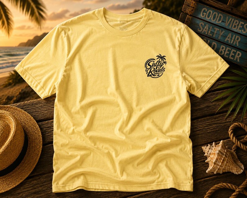 May include: A pale yellow short-sleeve t-shirt featuring a black circular graphic on the left chest. The graphic depicts a palm tree and a starfish with the text Salty Roads written in a stylized script font across the center of the circle.