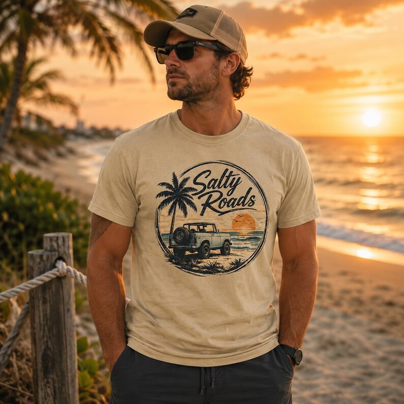 May include: A beige short-sleeve t-shirt featuring a circular graphic print on the chest. The design depicts a palm tree, a vintage off-road vehicle, and a sunset over the ocean with the text Salty Roads written in a script font.