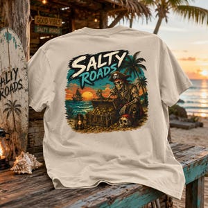 Puede incluir: Una camiseta beige con un estampado gr&aacute;fico de un esqueleto pirata con sombrero, sentado junto a un cofre del tesoro en una playa al atardecer. El dise&ntilde;o incluye palmeras, un barco en el agua, una linterna y el texto Salty Roads en negrita.