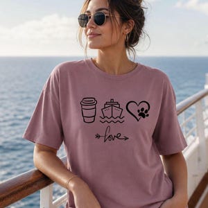 May include: Mauve-colored t-shirt with black graphic icons of a coffee cup, cruise ship, and heart with a paw print. The word "love" is written below the icons with an arrow. The shirt is worn by a person wearing sunglasses.