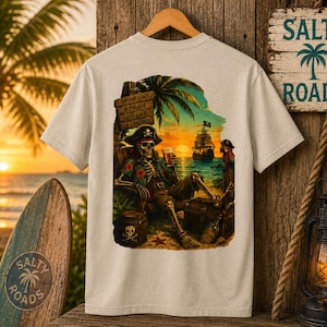 K&ouml;nnte beinhalten: Ein beiges T-Shirt mit einem Grafikdruck eines Skeletts mit Piratenhut und Blumenhemd, das bei Sonnenuntergang in einem Stuhl am Strand sitzt. Der Druck zeigt ein Schiff, einen Papagei, ein Fass und den Text: Drink like a pirate, live like a legend.