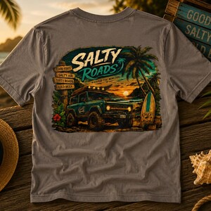 May include: A grey t-shirt featuring a graphic print of a teal off-road vehicle on a beach at sunset. The text reads Salty Roads, Chase the Horizon, Good Vibes, Salty Air, Dirty Roads, and Cold Beer. A palm tree and surfboard are also visible.