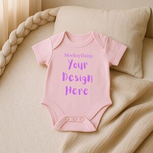Könnte beinhalten: Ein hellrosa Baby-Body mit kurzen Ärmeln und drei Druckknöpfen am unteren Saum. Der Text "MocksyDaisy" steht über den Worten "Your Design Here" in Lila. Der Body liegt auf einer beigen Oberfläche.