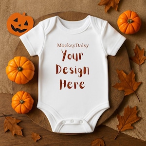 Könnte beinhalten: Ein weißer Baby-Body mit dem Text "MocksyDaisy Your Design Here" in Braun. Der Body ist umgeben von kleinen orangefarbenen Kürbissen, braunen Herbstblättern und einer geschnitzten Kürbisdekoration auf einer Holzoberfläche.