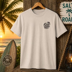 K&ouml;nnte beinhalten: Ein hellbeiges T-Shirt mit kurzen &Auml;rmeln und einer kleinen dunklen Grafik auf der linken Brustseite. Die Grafik zeigt ein Palmensymbol &uuml;ber dem Schriftzug Salty Roads in einer stilisierten Schreibschrift, erg&auml;nzt durch einen kleinen Stern.