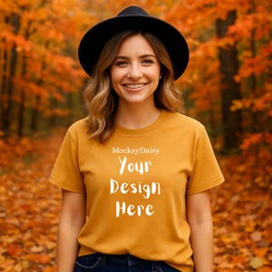 Könnte beinhalten: Senfgelbes T-Shirt mit dem weißen Text "Your Design Here". Das Model trägt einen schwarzen Hut. Der Hintergrund ist ein verschwommener Herbstwald mit orangefarbenen und roten Blättern.