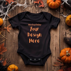 Könnte beinhalten: Schwarzer Baby-Body mit dem Text "Your Design Here" in Orange. Der Body ist von Kürbissen, Blättern und Spinnweben auf einem Holzgrund umgeben. Der Markenname "MocksyDaisy" steht über dem Text.