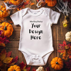 Könnte beinhalten: Weißer Baby-Body mit dem schwarzen Text "Your Design Here", umgeben von Kürbissen, Herbstblättern und Spinnweben auf einem Holzgrund. Der Body ist kurzärmlig und hat Druckknöpfe.