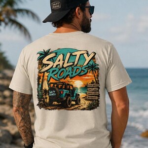 May include: A beige short-sleeve t-shirt featuring a graphic on the back of a teal off-road vehicle on a beach at sunset. The text reads SALTY ROADS, OCEAN BREEZE, SALTY AIR, GOOD VIBES, and NO HUMMIES. A surfboard is attached to the vehicle.