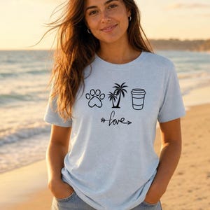 May include: Pale blue t-shirt with a paw print, palm tree, and coffee cup design. The word "love" is written below the graphics with an arrow. The shirt is made of a soft material.