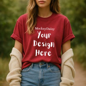 Könnte beinhalten: Ein weinrotes T-Shirt mit hochgekrempelten Ärmeln und dem Text "Your Design Here" in Weiß. Der Markenname "MocksyDaisy" steht über dem Text. Das Model trägt eine blaue Jeans und einen cremefarbenen Pullover.