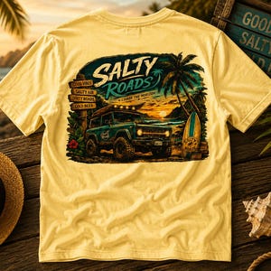 May include: A pale yellow t-shirt featuring a graphic on the back of a teal off-road vehicle parked on a beach at sunset. The design includes palm trees, a surfboard, and text that reads Salty Roads, Chase the Horizon, and Good Vibes.