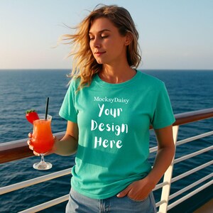 Könnte beinhalten: Türkisfarbenes T-Shirt mit dem Text "Your Design Here" und dem Markennamen "MocksyDaisy" in Weiß. Die Person trägt das T-Shirt und hält einen Cocktail mit Erdbeer-Garnitur. Der Hintergrund ist das Meer.