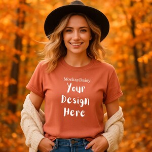 Puede incluir: Una camiseta de color óxido con el texto "Your Design Here" en blanco, bajo la marca "MocksyDaisy". La modelo lleva un sombrero negro y una rebeca color crema. El fondo es una vista borrosa del follaje otoñal.