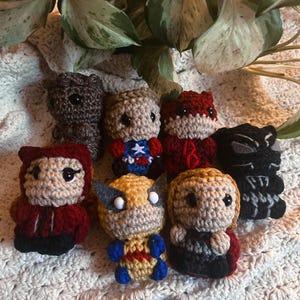 Puede incluir: Una colección de muñecos de superhéroes hechos a mano con ganchillo. Los muñecos presentan varios personajes con colores y diseños distintos, incluyendo rojo, negro, amarillo y azul. Están colocados sobre una superficie blanca de ganchillo.