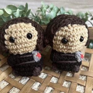 May include: Two handmade crochet dolls with tan faces, dark brown hair, and black eyes. They wear black outfits with gray and red accents. The dolls are displayed on a woven basket.