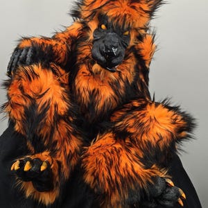 May include: A full-body creature costume featuring long, shaggy faux fur in vibrant orange and black. The headpiece has a black snout, pointed ears, and glowing yellow eyes. The hands and feet are black with prominent, sharp orange-tipped claws.