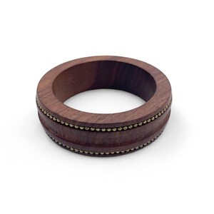 Puede incluir: Un brazalete de madera marrón oscuro con una banda de pequeñas cuentas doradas. El brazalete es circular y tiene un acabado liso y pulido. Las cuentas están espaciadas uniformemente alrededor del centro del brazalete.