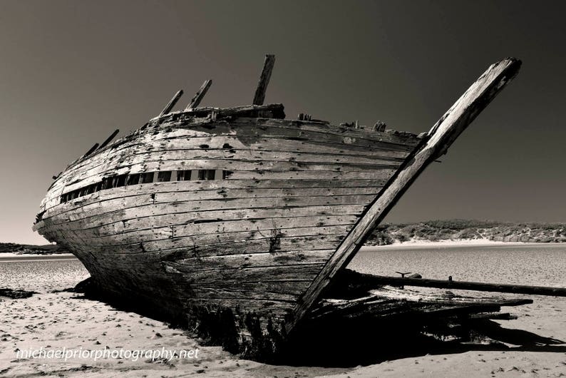 Peut inclure: Photographie en noir et blanc d'une &eacute;pave de navire en bois sur une plage de sable. La coque du navire est partiellement intacte, avec des planches et des &eacute;l&eacute;ments structurels visibles. L'image capture la texture vieillie du navire et le paysage environnant.