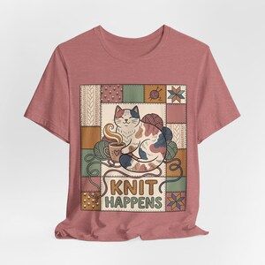 Può includere: Una t-shirt rosa polvere con una grafica centrale raffigurante un gatto calico che tiene una tazza fumante, circondato da gomitoli di lana e uno sfondo patchwork. Il testo KNIT HAPPENS è stampato in grassetto in stampatello sotto il disegno.