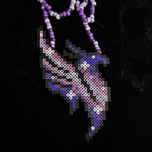 May include: A beaded necklace with a pendant in shades of purple, pink, and white. The pendant is a stylized feather or wing shape, made from small, colorful beads. The necklace itself is made of purple and white beads.