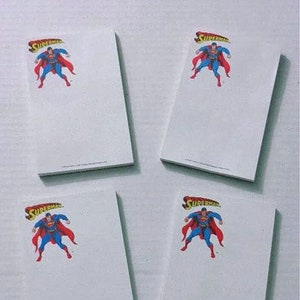May include: Four white notepads featuring the Superman logo and image. The logo is red and yellow, with the word "Superman" above a cartoon image of Superman in a blue suit and red cape.