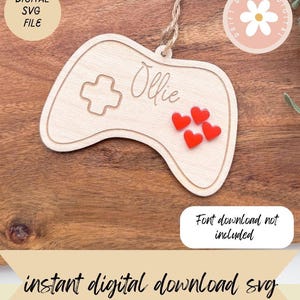 May include: A wooden game controller-shaped ornament with the name "Ollie" and red heart embellishments. It features a cross symbol and is suspended by twine. The text "DIGITAL SVG FILE" and "instant digital download svg" are also visible.