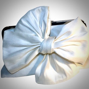 May include: A white satin clutch bag with a large, oversized bow. The bag has a silver metal frame.