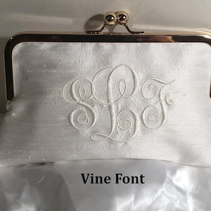 Puede incluir: Un bolso de mano tipo sobre de color blanco con un marco de metal dorado y cierre de boquilla. La parte frontal está bordada con un monograma grande en color blanco con las letras S, L y F en cursiva. El texto Vine Font es visible.