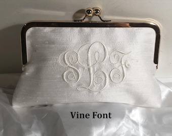 May include: A white clutch bag with a gold-toned metal frame and a kiss-lock closure. The fabric features a subtle sheen and is embroidered with a large, white, cursive monogram featuring the letters S, L, and F. The text Vine Font is visible.