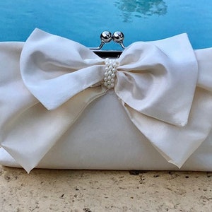 May include: A white satin clutch bag with a large bow and pearl embellishments. The bag has a silver clasp.