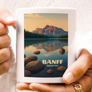 May include: White ceramic mug featuring a scenic illustration of Banff National Park. The design includes a mountain range, lake, and the text "BANFF National Park" in a dark blue font. The mug is held by a person wearing gold rings.