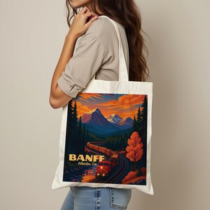 May include: Cream-colored tote bag featuring a colorful illustration of a train winding through a mountain landscape at sunset. The word "BANFF" and "Alberta, Ca" are printed on the bag.