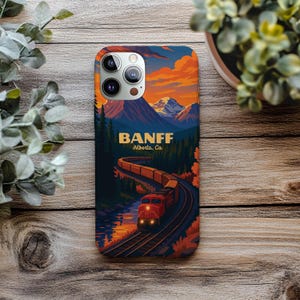 May include: A phone case featuring a scenic illustration of Banff, Alberta, with a train winding through a forest and mountains under a sunset sky. The word "BANFF" is prominently displayed, along with "Alberta, Ca."