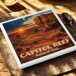 May include: A square, white-framed art print featuring a landscape of Capitol Reef National Park at sunset. The artwork depicts a desert scene with red rock formations, sparse vegetation, and a brilliant sun. The text "CAPITOL REEF National Park" is printed on the bottom.