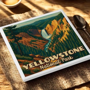 May include: A square coaster with a scenic illustration of Yellowstone National Park. The artwork features a waterfall, river, and forest, with the words "YELLOWSTONE National Park" in a serif font. The coaster is on a textured surface.
