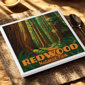 May include: A square coaster with a Redwood National Park design. The image features tall redwood trees, green ferns, and the words "REDWOOD National Park" in orange. The coaster is on a textured, tan surface.
