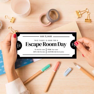 May include: A black and white "Escape Room Day" ticket held by a person. The ticket reads "Save the Date" and includes fields for the recipient's name, date, and location. Other items on the wooden surface include pens, tape, and paper clips.