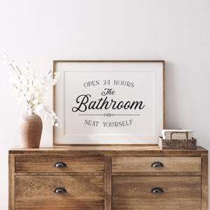 May include: A framed bathroom sign with the text "OPEN 24 HOURS The Bathroom SEAT YOURSELF" in black script. The sign is displayed on a wooden dresser with a vase of white flowers and a small woven basket.