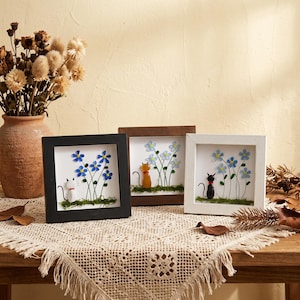 May include: Three framed art pieces featuring cat figures among blue flowers. The frames are black, brown, and white. The cats are depicted in various colors, with green moss at the base of the flowers. The artwork is displayed on a wooden table.