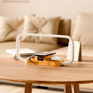 May include: A wooden charging station with a digital clock displaying 22:27. It holds a phone, smartwatch, and wireless earbuds. The station has the name "Michelle" engraved on it, and is on a wooden table.